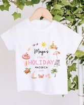 A white t-shirt with a printed design featuring various summer-themed illustrations, including a beach, palm tree, umbrella, and other beach-related objects. The text on the t-shirt reads "Maya's FIRST HOLIDAY MAJORCA 2023".