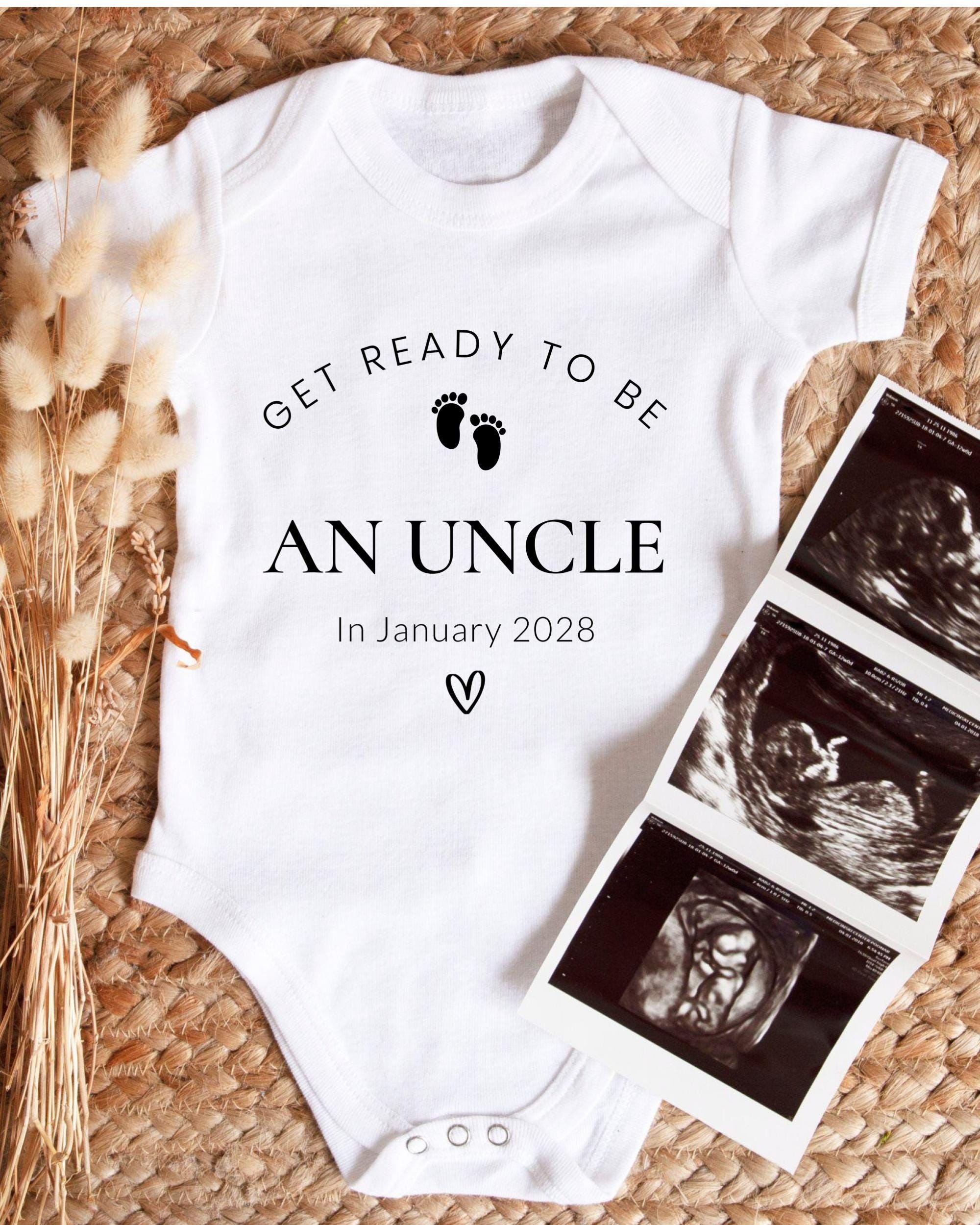 A white baby onesie with the text "GET READY TO BE AN UNCLE In January 2028" printed on it, along with a pair of baby footprints. The onesie is placed next to a set of three black and white baby ultrasound images.