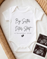 A white baby onesie with the text "Big Sister Duties Start in November 2026" printed on it, along with a pair of black and white baby footprints. The onesie is placed on a white blanket, and there are two black and white baby images next to it.