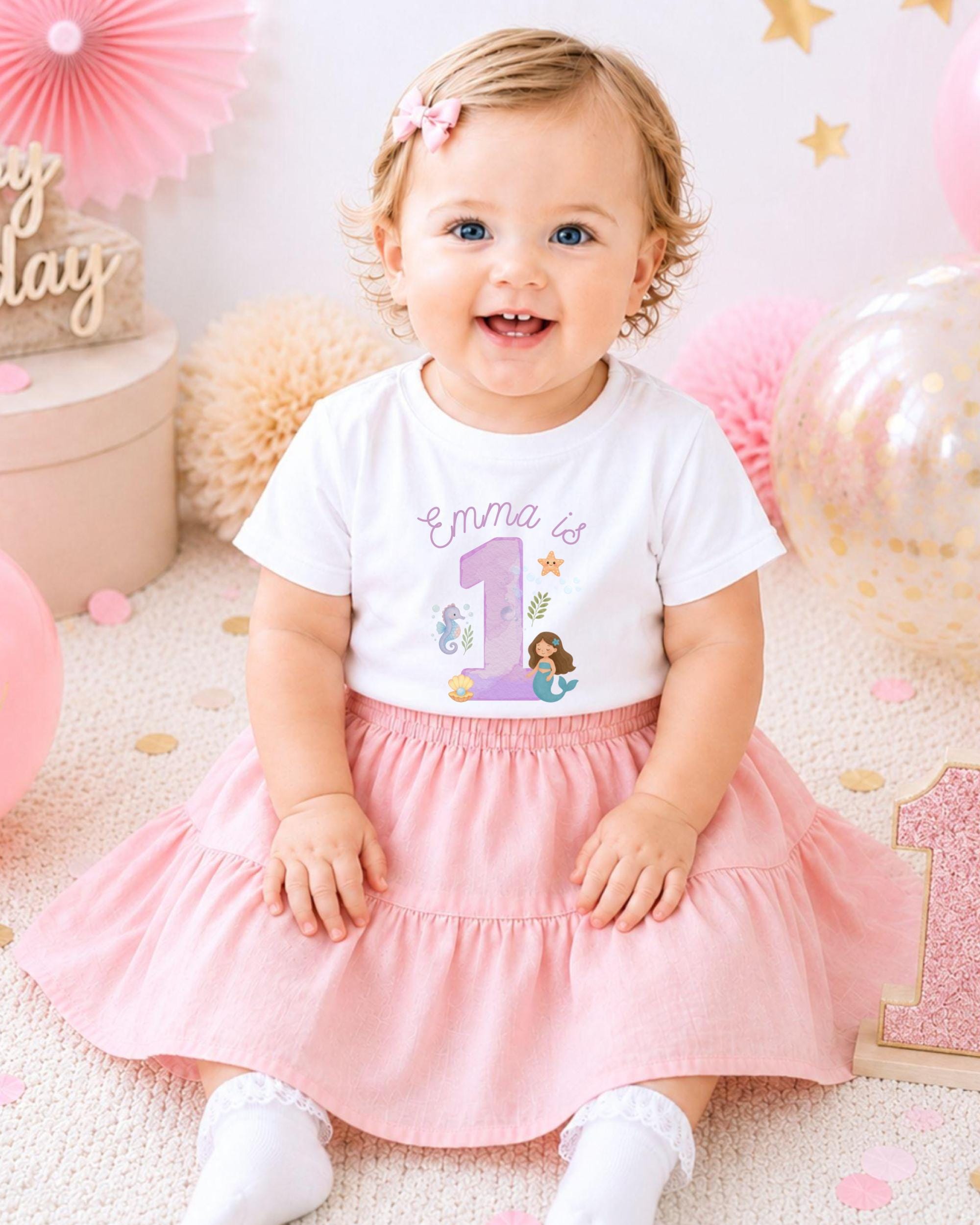 A smiling baby girl wearing a white t-shirt with a mermaid design and a pink skirt, sitting on a pink and white background with balloons and confetti.