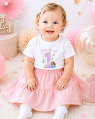 A smiling baby girl wearing a white t-shirt with a mermaid design and a pink skirt, sitting on a pink and white background with balloons and confetti.
