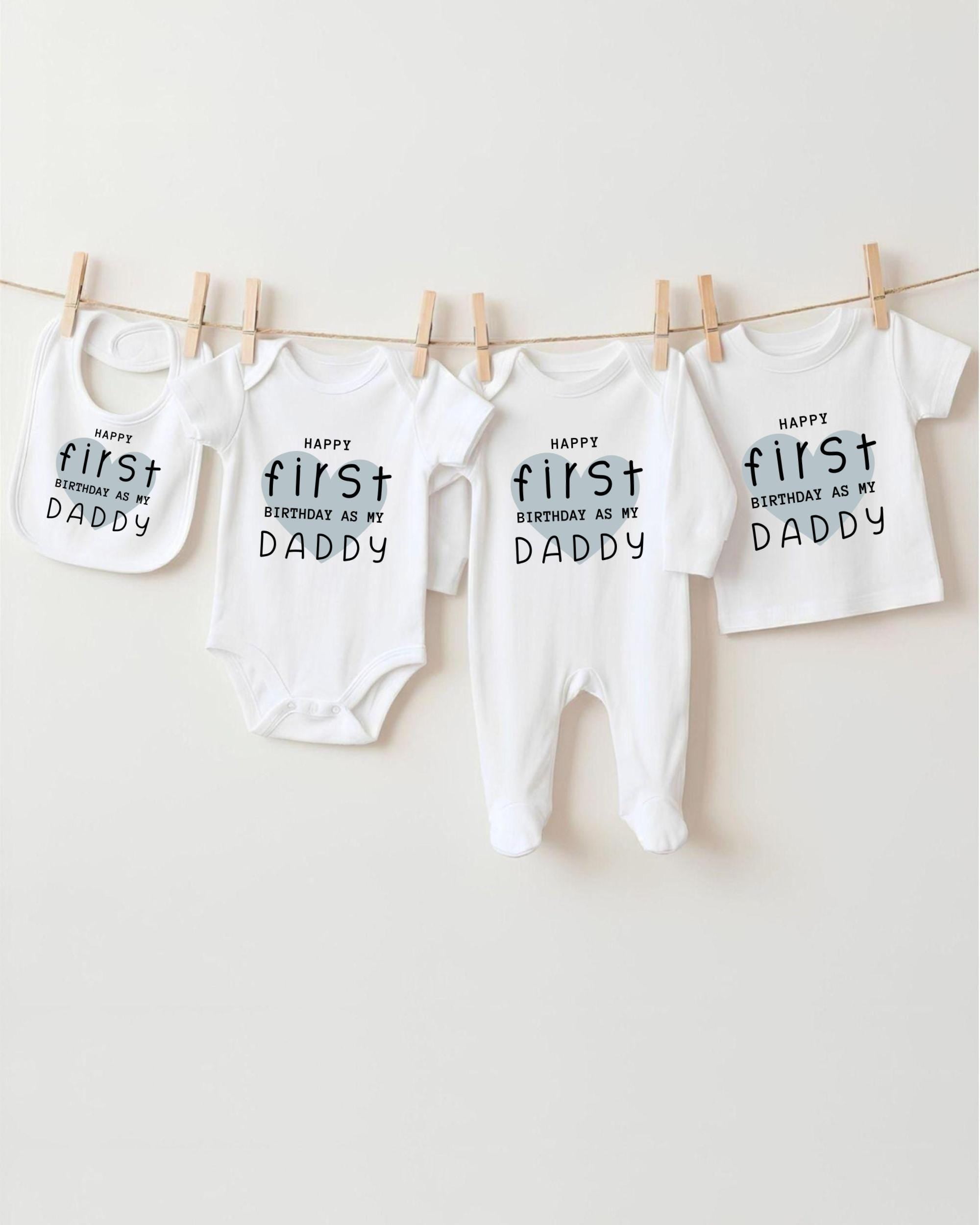 A row of white baby clothes with the text "Happy first birthday as my Daddy" printed on them, hanging on a clothesline.
