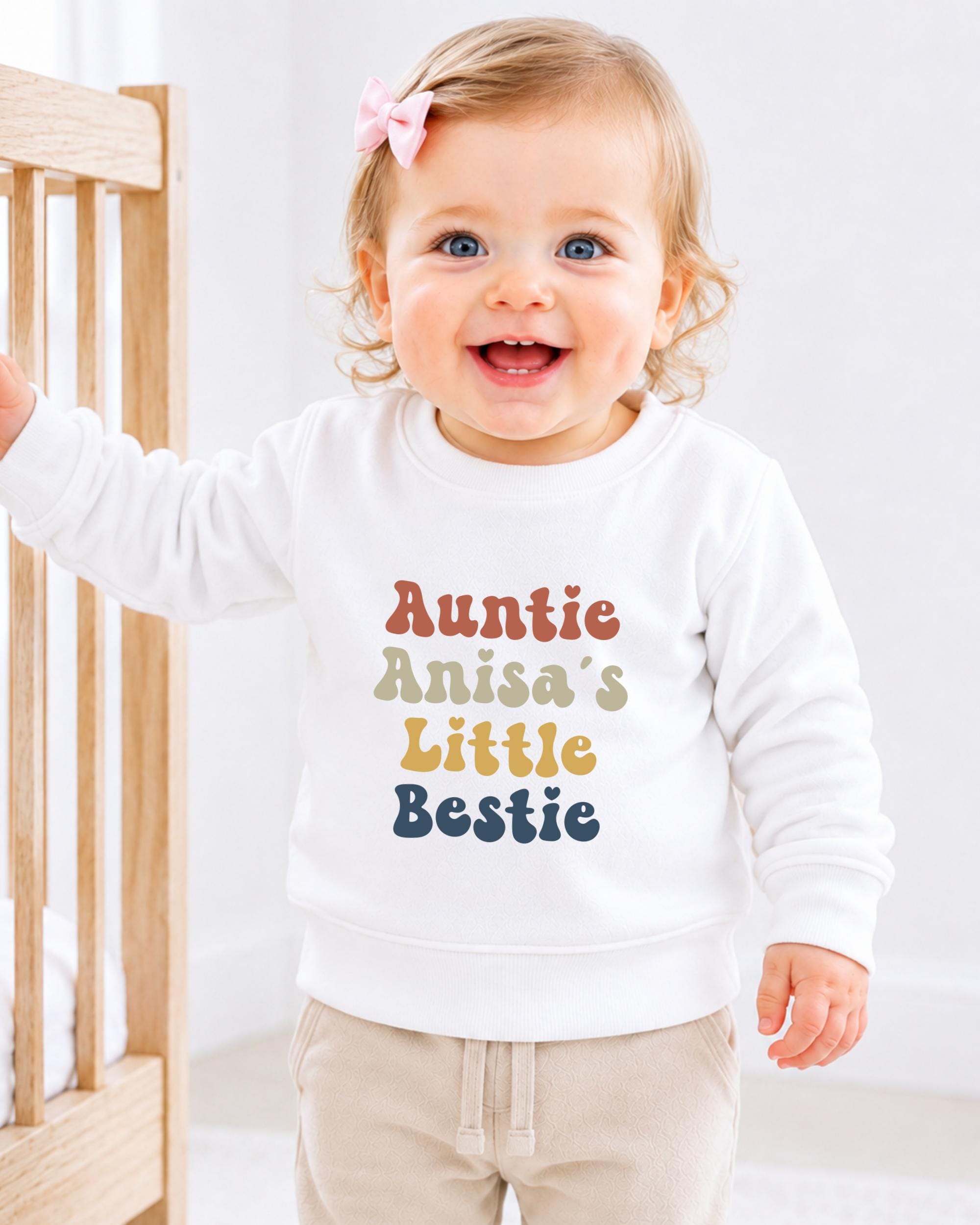 A smiling baby girl wearing a white sweatshirt with the text "Auntie Anisa's Little Bestie" printed on it.