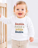 A smiling baby girl wearing a white sweatshirt with the text "Auntie Anisa's Little Bestie" printed on it.