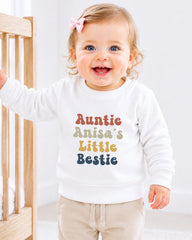A smiling baby girl wearing a white sweatshirt with the text "Auntie Anisa's Little Bestie" printed on it.