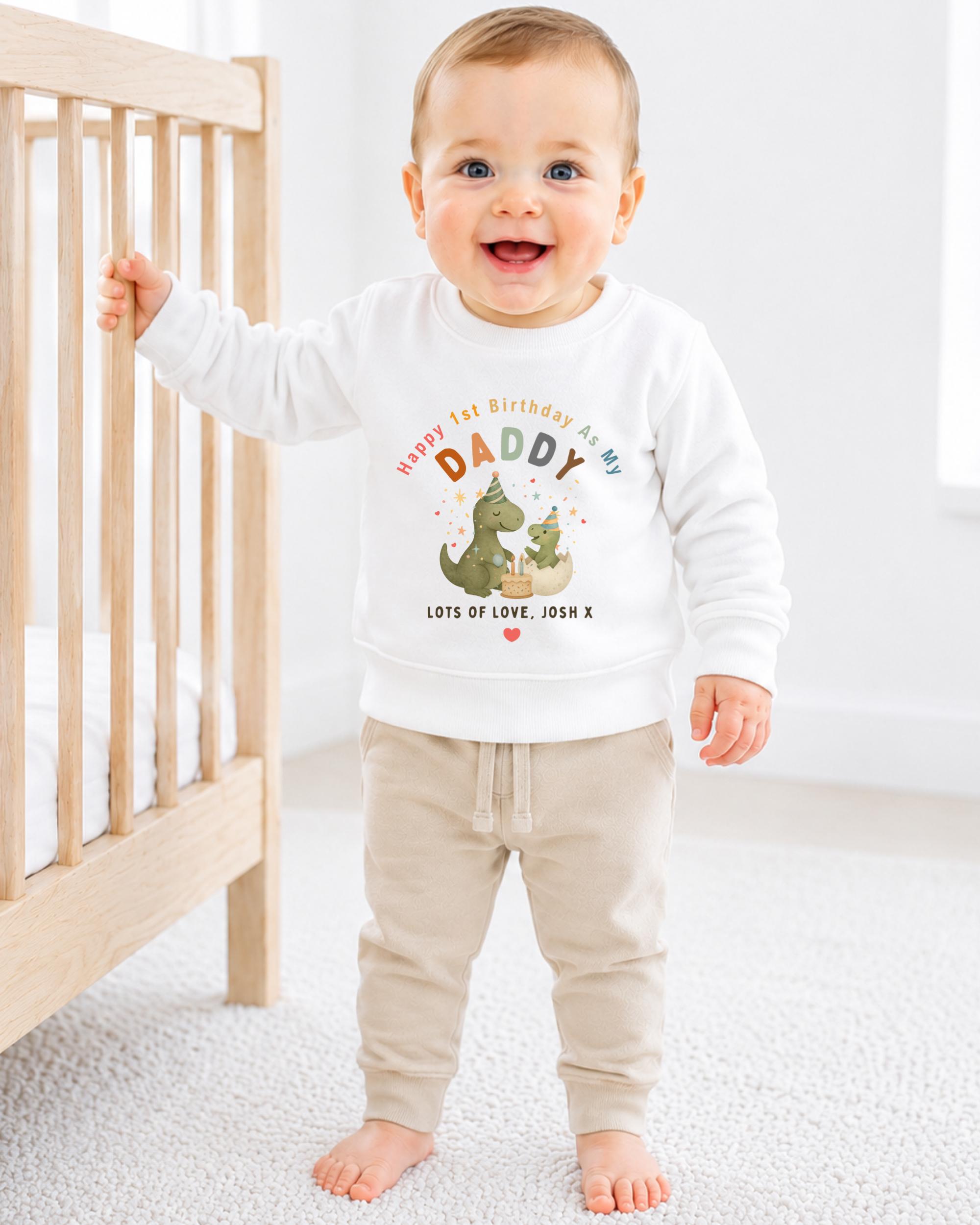 A smiling baby boy wearing a white sweatshirt with a dinosaur graphic and text that says "Happy 1st Birthday As My Daddy Lots of Love Josh X".