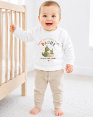 A smiling baby boy wearing a white sweatshirt with a dinosaur graphic and text that says "Happy 1st Birthday As My Daddy Lots of Love Josh X".