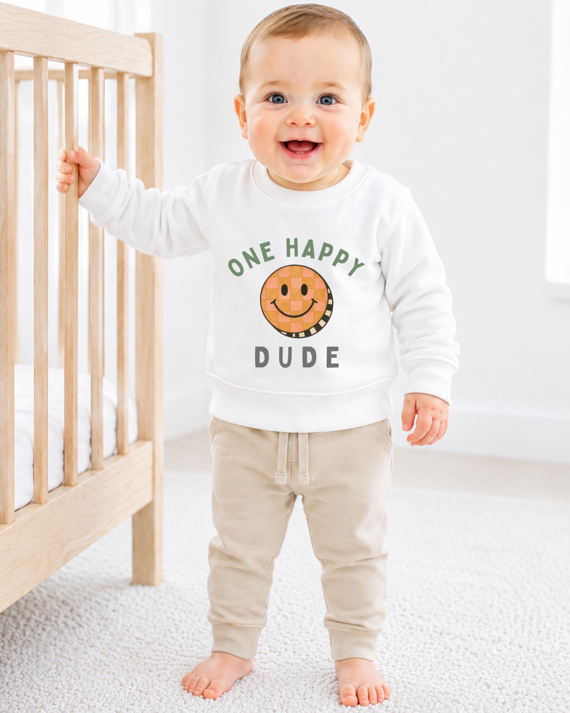 A smiling baby boy wearing a white sweatshirt with a smiley face and the text "One happy dude" is standing next to a wooden crib.