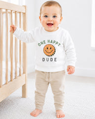 A smiling baby boy wearing a white sweatshirt with a smiley face and the text "One happy dude" is standing next to a wooden crib.