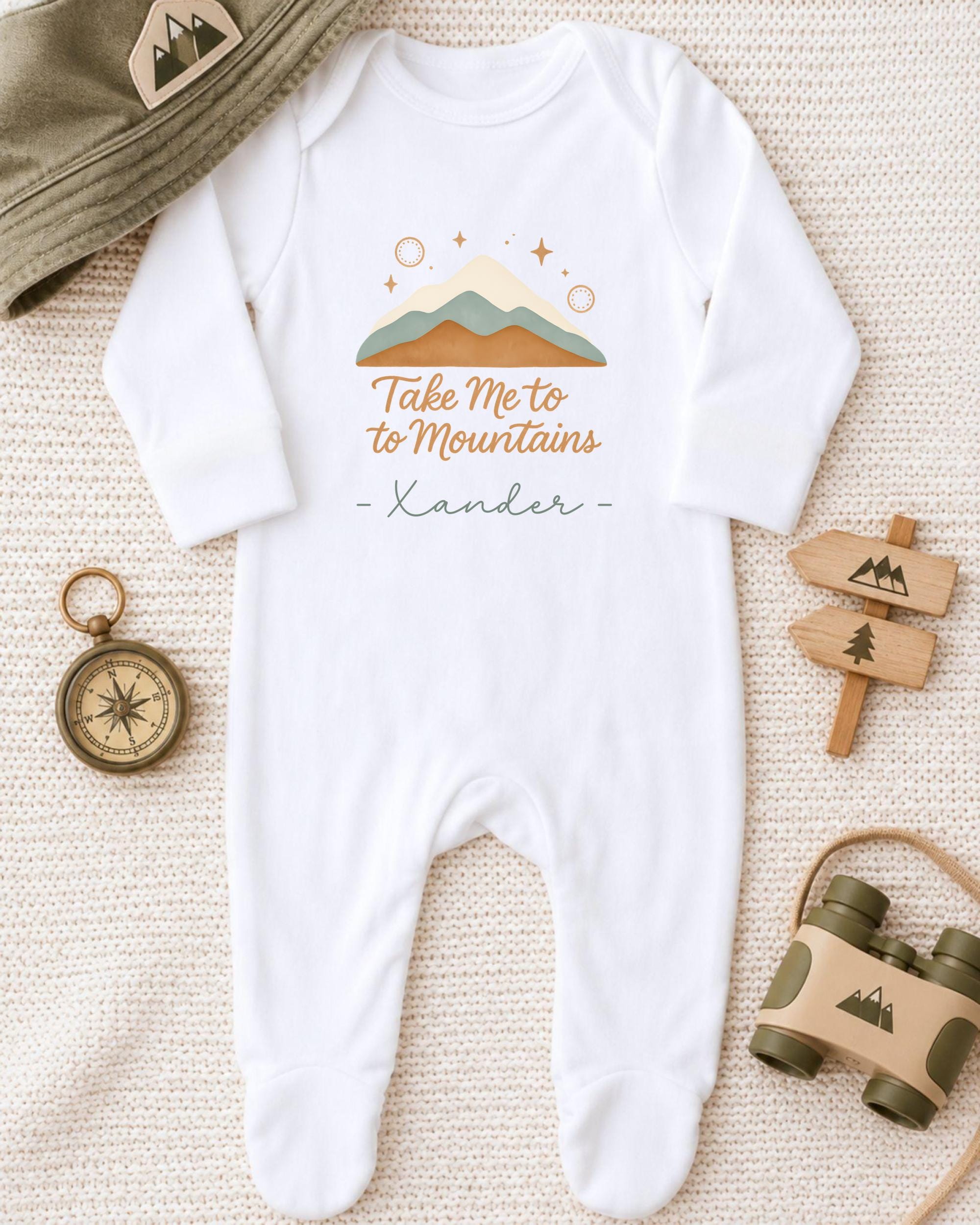 A white onesie with a mountain and star design, along with a compass, a hat, and a toy binoculars, all arranged on a beige surface.