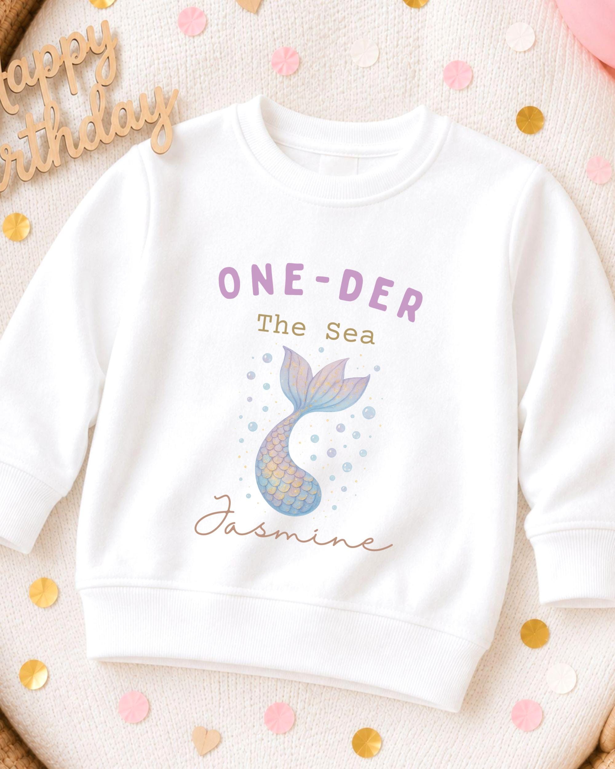 A white sweatshirt with a mermaid tail design and the text "ONE-DER The Sea Jasmine" printed on it, along with a "Happy Birthday" sign.