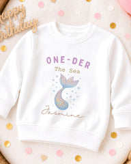 A white sweatshirt with a mermaid tail design and the text "ONE-DER The Sea Jasmine" printed on it, along with a "Happy Birthday" sign.