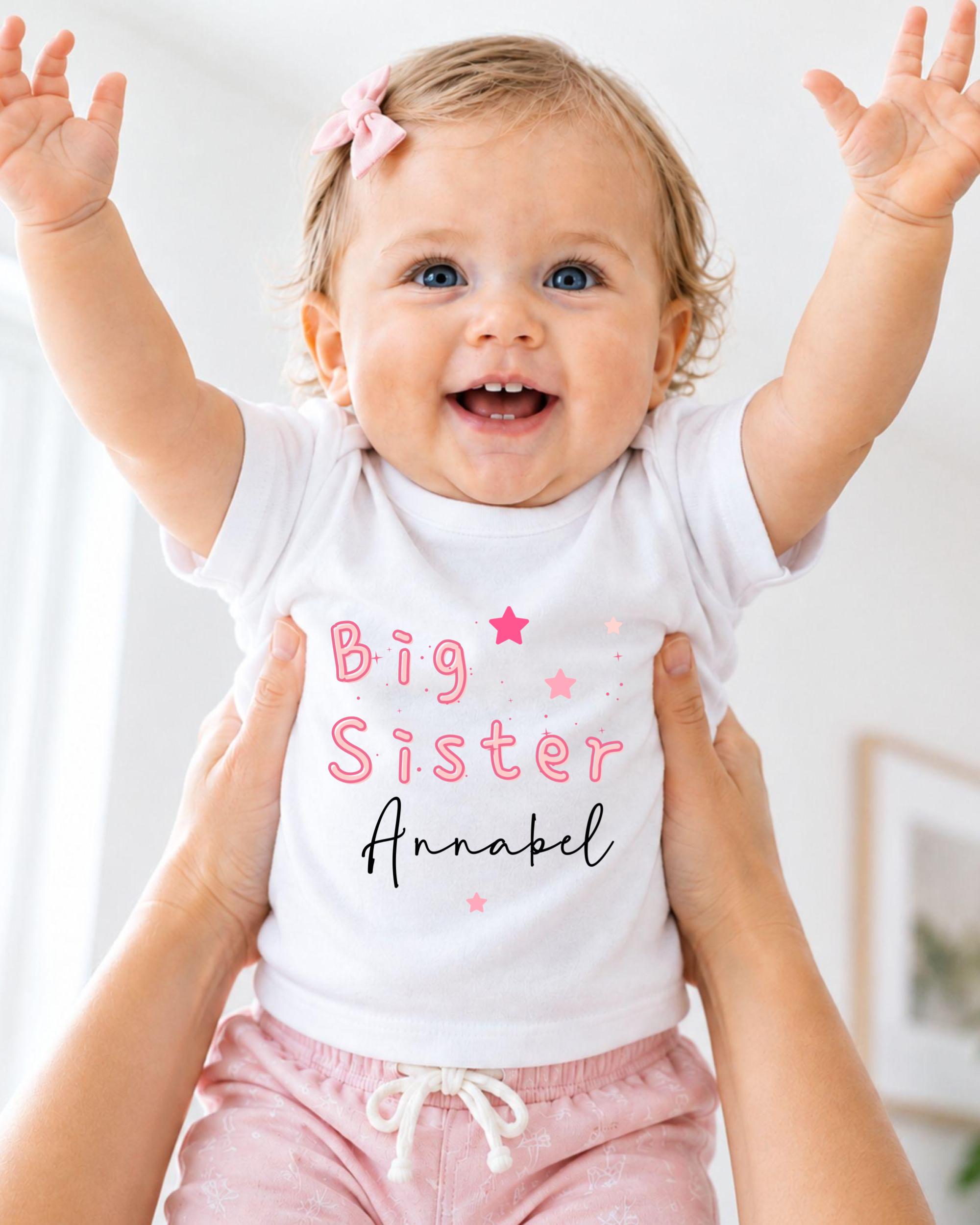 A smiling baby girl wearing a white shirt with the text "Big Sister Annabel" is being held up by someone's arms.