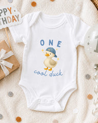 A white onesie with a cartoon duck and the text "One cool duck" printed on it, along with a "Happy Birthday" sign and a balloon.