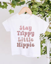 A white t-shirt with the text "Stay Trippy Little Hippie" printed on it, hanging on a wooden hanger.