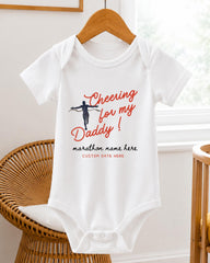 A white baby onesie with a graphic design and text printed on it, hanging on a wooden hanger.