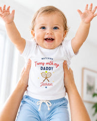 A smiling baby wearing a white t-shirt with a tennis racket and the text "Watching Tennis with my Daddy Jackson" is being held up by someone's arms.