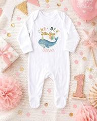 A white onesie with a whale design and the text "One-DER The Sea Harper" printed on it, surrounded by birthday decorations and confetti.
