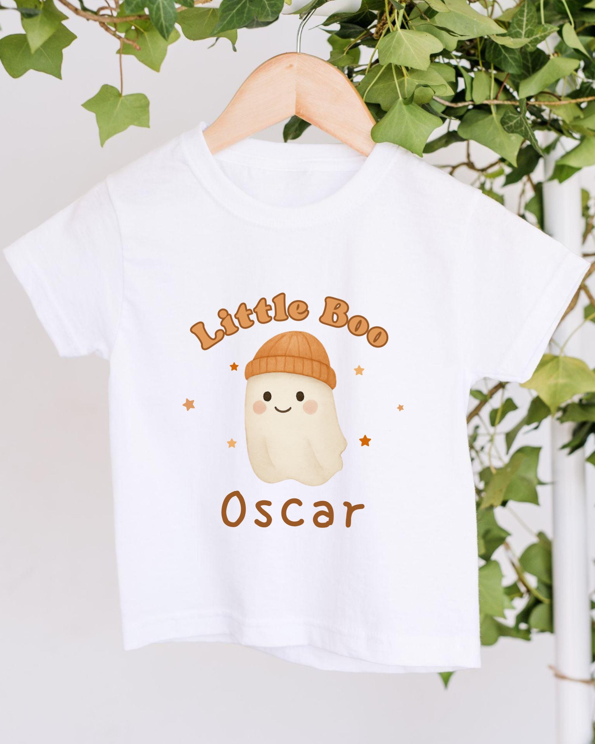 A white t-shirt with a cartoon ghost wearing a hat and the text "Little Boo Oscar" printed on it, hanging on a wooden hanger against a backdrop of greenery.