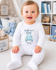 A smiling baby boy wearing a white onesie with a blue car design and the text "Halfway to One Scoop" is sitting on the floor in a nursery setting.