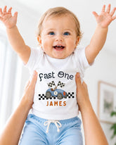 A smiling baby wearing a white t-shirt with a racing car graphic and the name "James" printed on it, being held up by someone's arms.