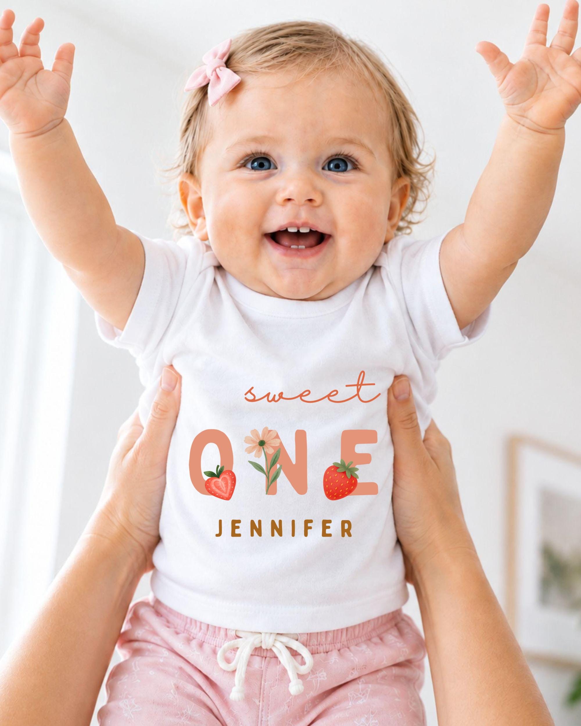 A smiling baby girl wearing a white t-shirt with the text "Sweet One Jennifer" and pink shorts, being held up by someone's arms.