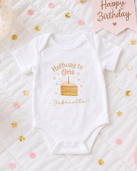 A white onesie with a birthday cake design and the text "Halfway to One Gabriella" printed on it, surrounded by confetti and a "Happy Birthday" sign.