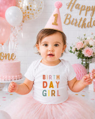 A young girl wearing a birthday hat and a white t-shirt with the text "Birthday Girl" is posing in front of a birthday cake and balloons.