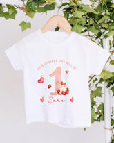 A white t-shirt with a personalized design hanging on a wooden hanger, featuring a pink number one, strawberries, and hearts.