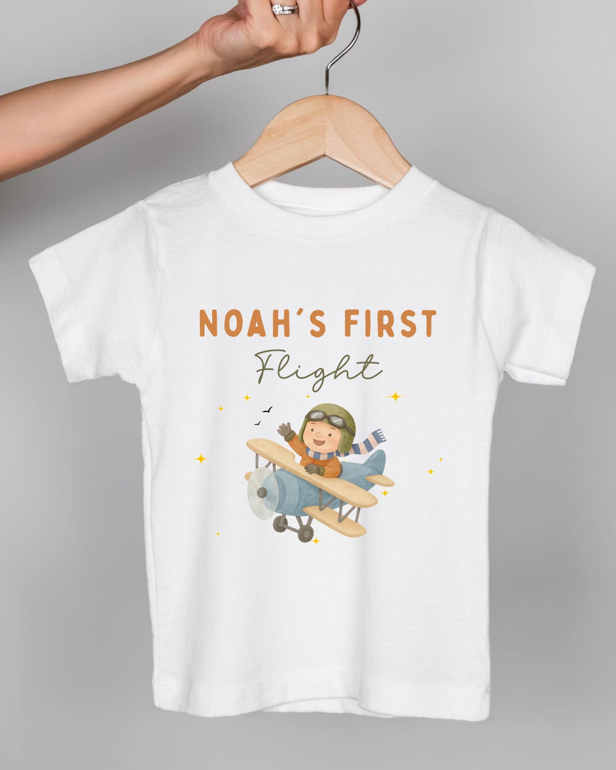 A white t-shirt with a cartoon image of a boy in a pilot's suit and helmet, along with the text "Noah's first flight" printed on it.