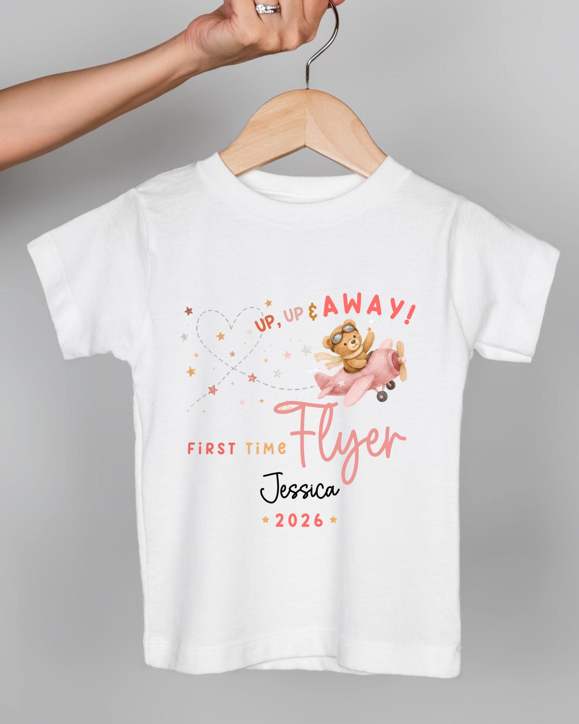 A white t-shirt with a graphic design featuring a cartoon character and text that says "Up, up & away! First time flyer Jessica 2026".