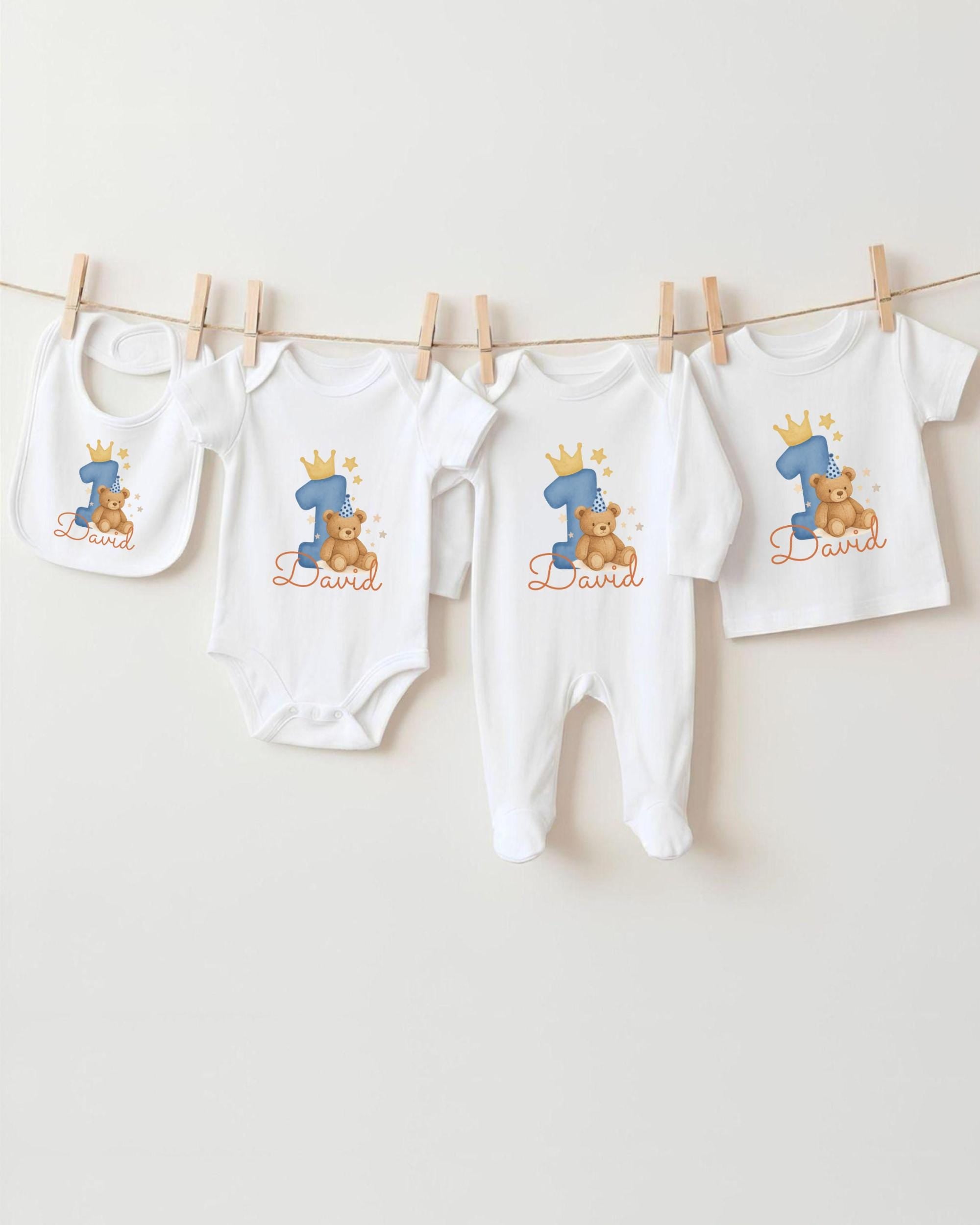 A collection of white baby clothes, including onesies and pants, with a blue number one on each, hanging on a clothesline.