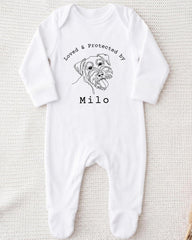 A white onesie with a black line drawing of a dog's face and the text "Loved & Protected by" above it, along with the name "Milo" below the drawing.