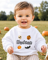 A smiling baby boy wearing a white sweatshirt with a Halloween-themed design, including a skull, pumpkin, bat, and pumpkin.