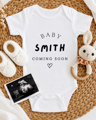 A baby announcement t-shirt with the text "BABY SMITH COMING SOON" printed on it, along with a pair of white crocheted baby booties and a crocheted rabbit toy.