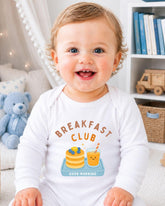 A smiling baby wearing a white onesie with a breakfast club graphic and text.