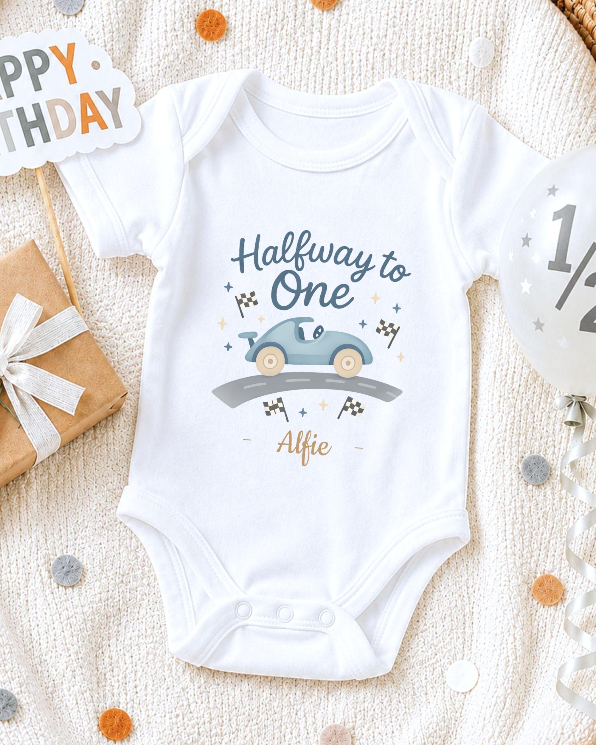 A white onesie with a car graphic and the text "Halfway to One" printed on it, along with the name "Alfie" below the car. The onesie is laid out on a white blanket, surrounded by confetti and a birthday card.
