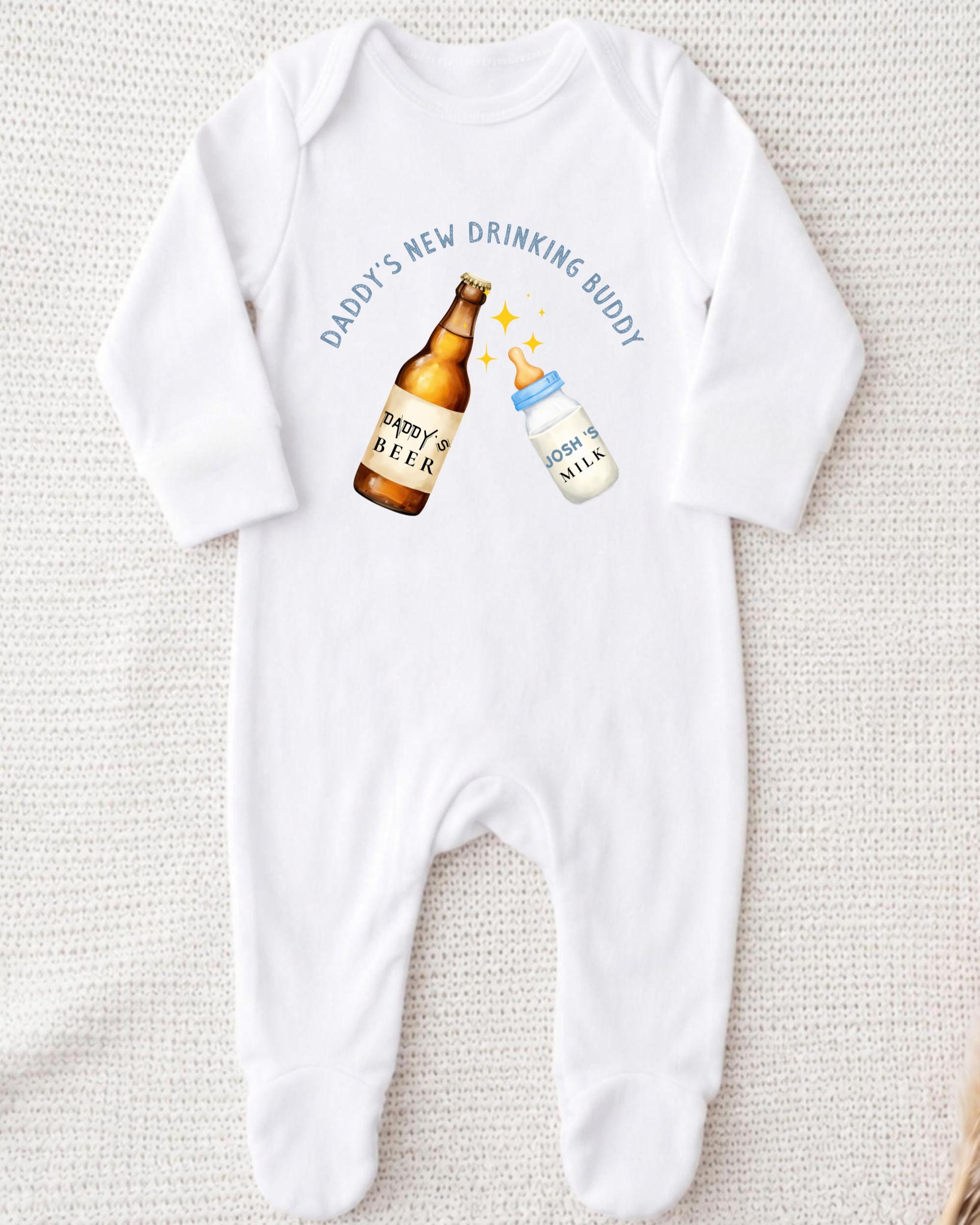 A white onesie with a graphic design on it, depicting a beer bottle and a baby bottle. The text above the graphic says "DADMY'S NEW DRINKING BUDDY".