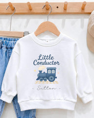 A white sweatshirt with a train design and the text "Little Conductor Sutton" printed on it, hanging on a wooden hanger.