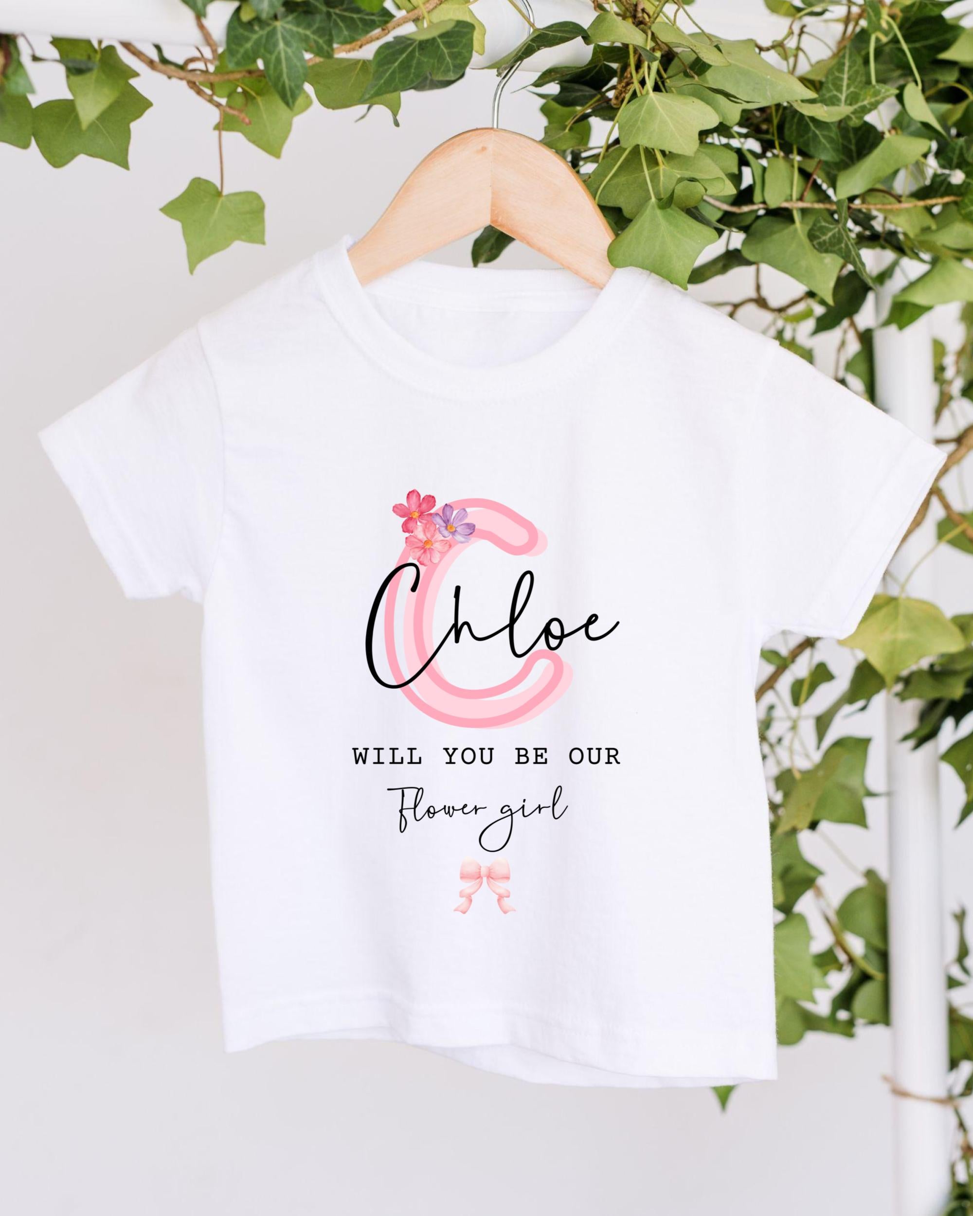 A white t-shirt with a pink floral design and text printed on it, hanging on a wooden hanger.