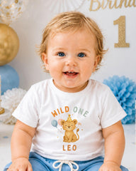 A smiling baby boy wearing a white t-shirt with a cartoon lion and the text "Wild one Leo" is sitting in front of a birthday party backdrop.