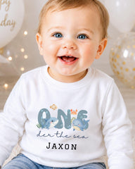 A smiling baby boy wearing a white shirt with a cartoon shark and the text "ONE der the sea JAXON" printed on it.