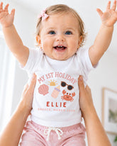 A smiling baby girl wearing a white t-shirt with a graphic design and text, being held up by someone's arms.