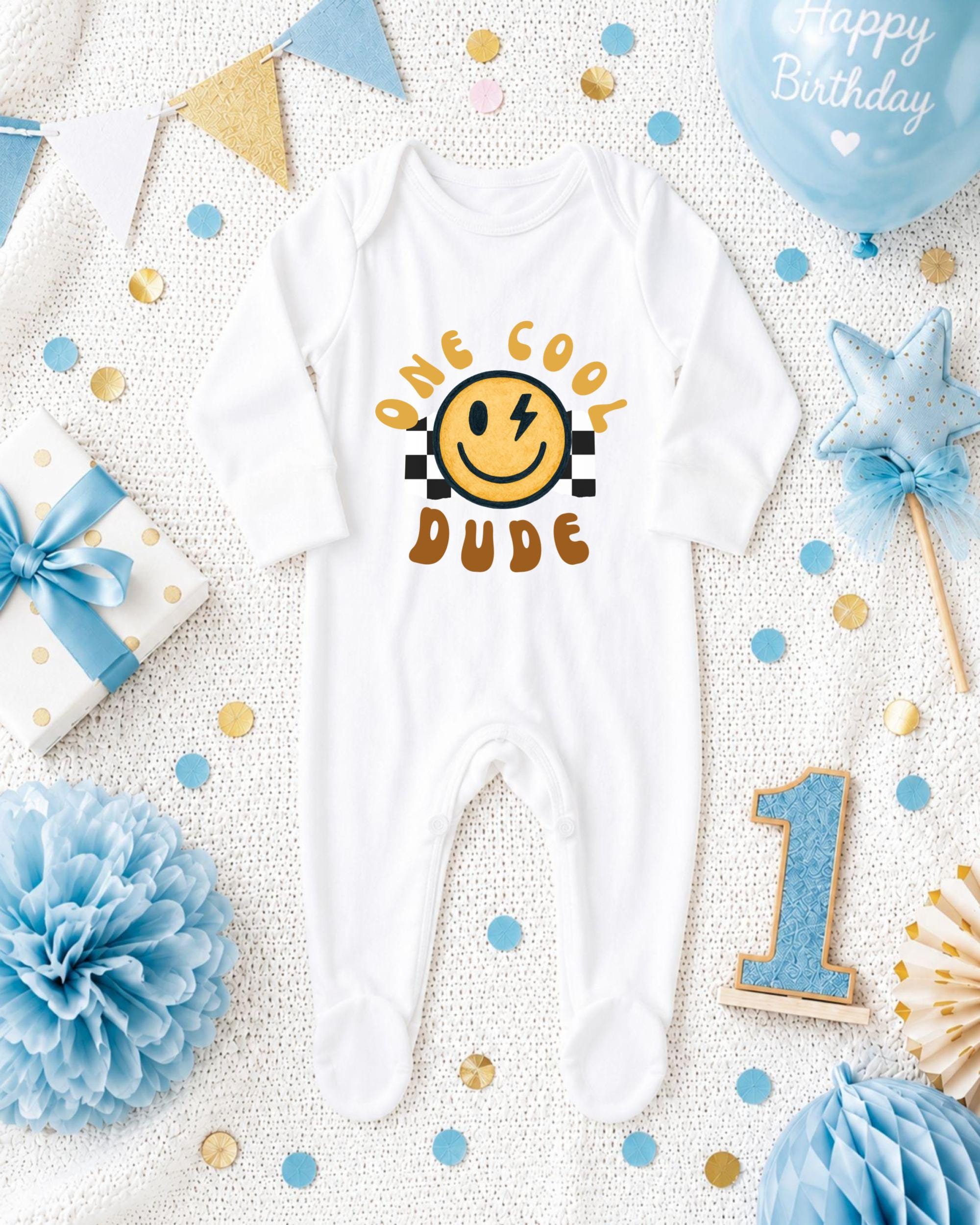 A white onesie with a smiley face and the text "One cool dude" printed on it, surrounded by birthday decorations and a blue balloon.