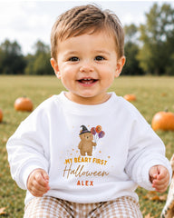 A smiling baby boy wearing a white sweatshirt with a Halloween-themed design and text.