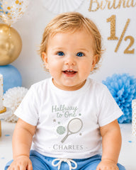A smiling baby boy wearing a white t-shirt with a tennis racket and the text "Halfway to One" is sitting in front of a birthday party backdrop.