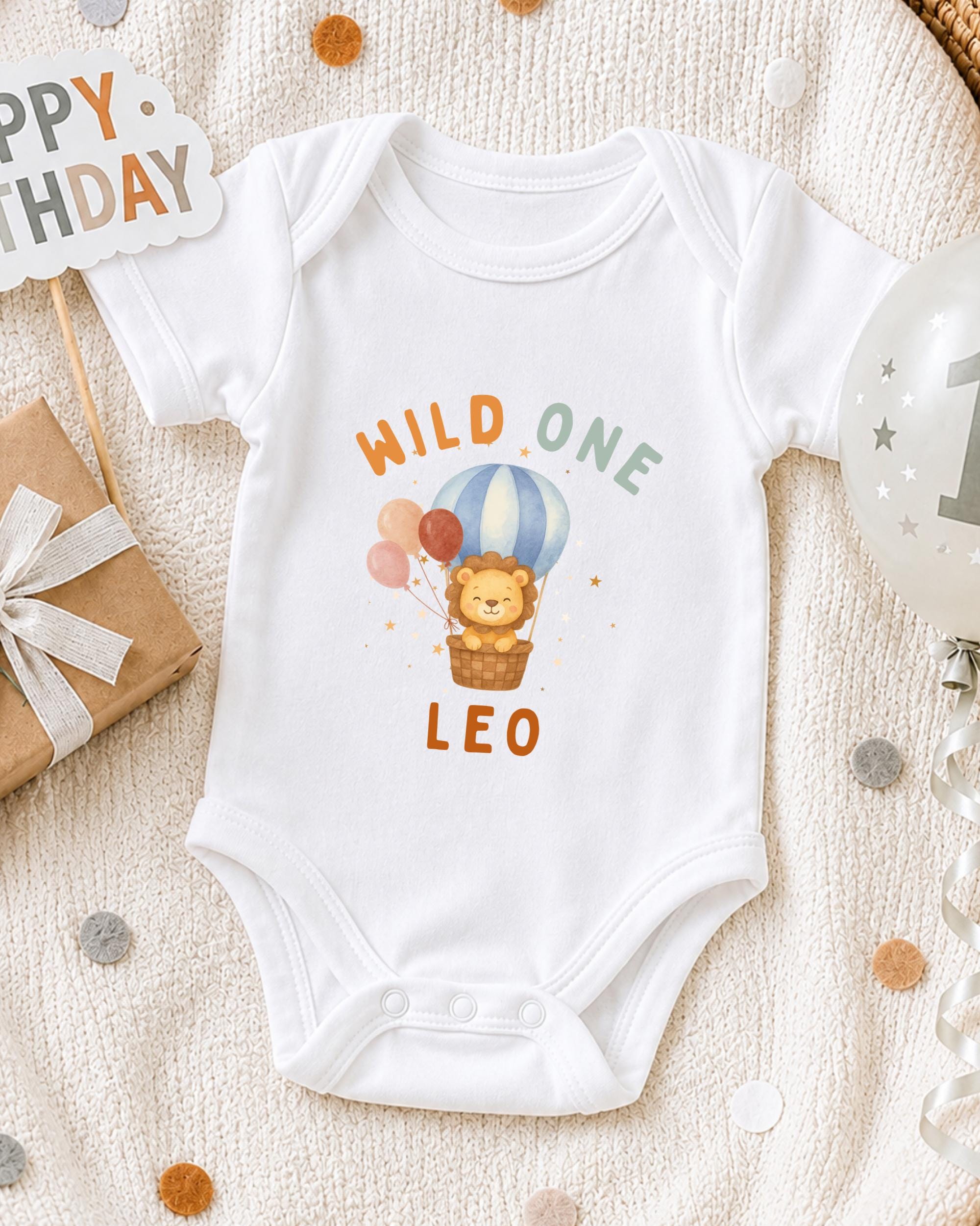 A white onesie with a cartoon lion and a hot air balloon design, along with a "Happy Birthday Leo" sign, is displayed on a white blanket.