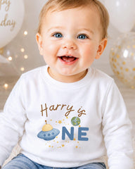 A smiling baby boy wearing a white shirt with a graphic and text printed on it.