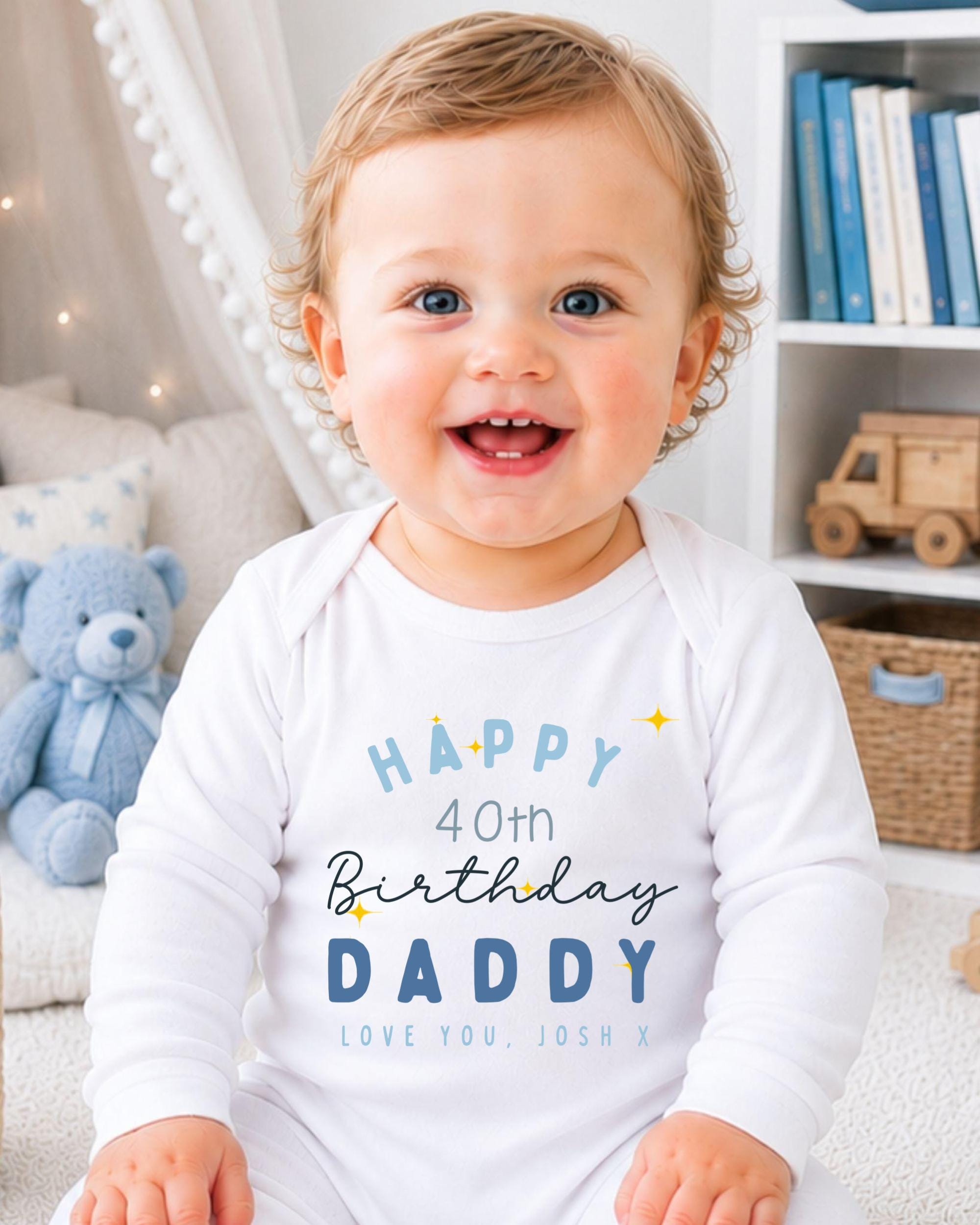 A smiling baby boy wearing a white onesie with text that says "Happy 40th Birthday Daddy Love You Josh X" is sitting in a room with a teddy bear and bookshelf in the background.