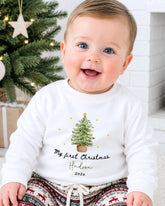 A smiling baby wearing a white sweater with a Christmas tree design and text that says "My first Christmas Hudson 2020x".